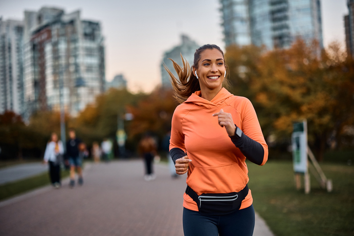 A woman in an orange hoodie is jogging outdoors with a smile with city buildings in the background.