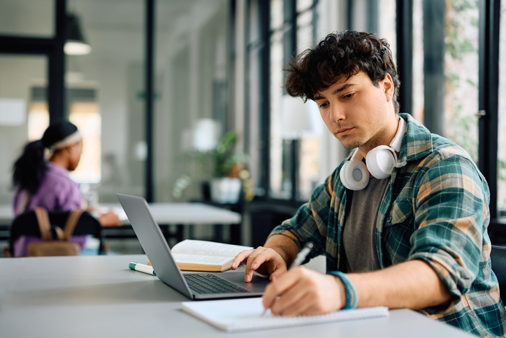 A focused student with headphones around his neck is seated at a desk, viewing course work on his laptop while simultaneously writing in a notebook.