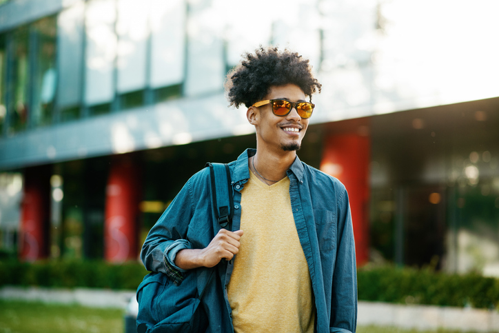 A smiling young man with sunglasses, wearing a denim shirt over a yellow t-shirt and carrying a school bag over his shoulder