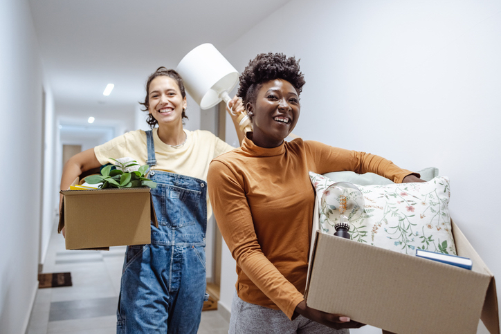 Two smiling female students, one with overalls and carrying a box with a plant, and the other with a turtleneck shirt carrying a box with a pillow and book, moving into their new dorm.