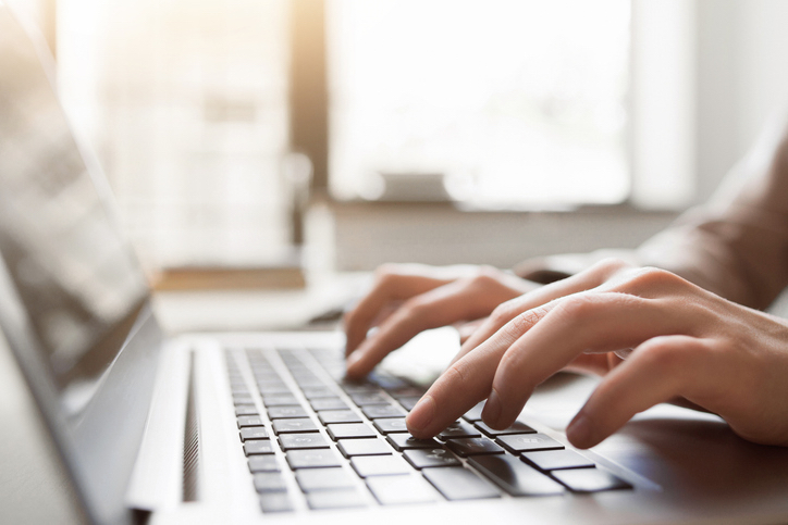 Close-up of a person's hands typing on a laptop keyboard, with a bright window in the background
