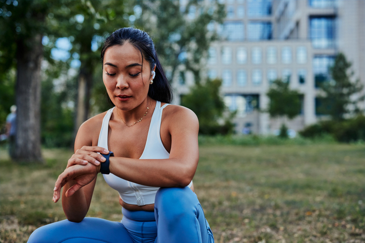 A woman in athletic wear resting outdoors in a park, looking down at a smartwatch on her wrist.