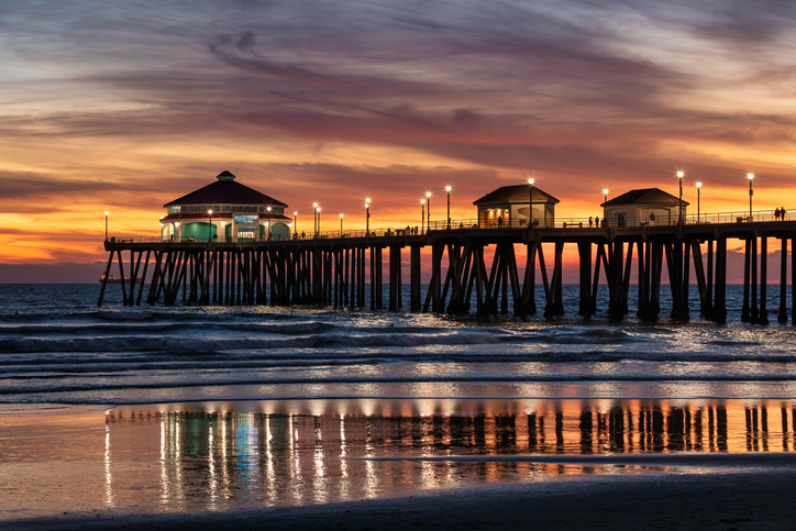 A colorful sunset over the ocean, illuminating the Huntington Beach Pier. The sky is filled with orange and purple clouds, and the lights of the pier reflect on the gentle waves and wet sand below.