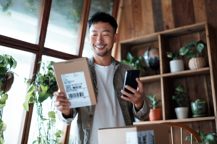 A smiling man in a casual jacket stands in his modern home, looking at his smartphone while holding a cardboard package.