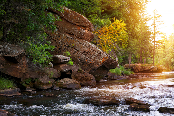 A sunlit forest river flowing past large, moss-covered rocks on the left. Golden sunlight streams through the trees on the right, illuminating the water.