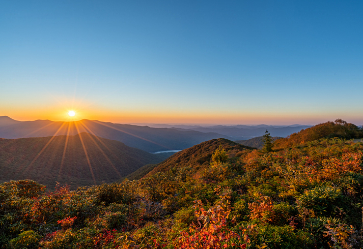 The fiery colors of a sunset wash over rolling hills, lighting up a dense forest of autumn foliage.